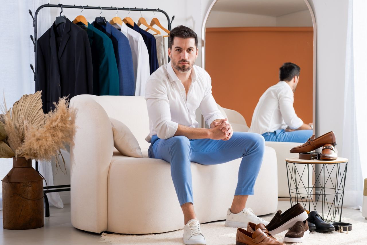 Contact A fashionable man in a modern showroom with elegant outfits and shoes displayed around him.